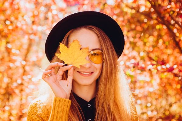 Moça bonita usando óculos de sol e chapéu, em meio a uma paisagem de outono, segurando uma folha de árvore.